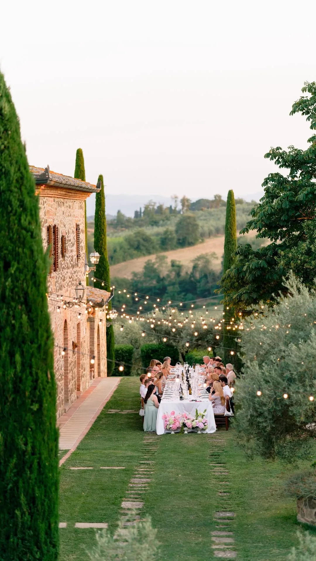 Lunga tavolata di un matrimonio sotto le luci a filo, tra cipressi e ulivi, al tramonto