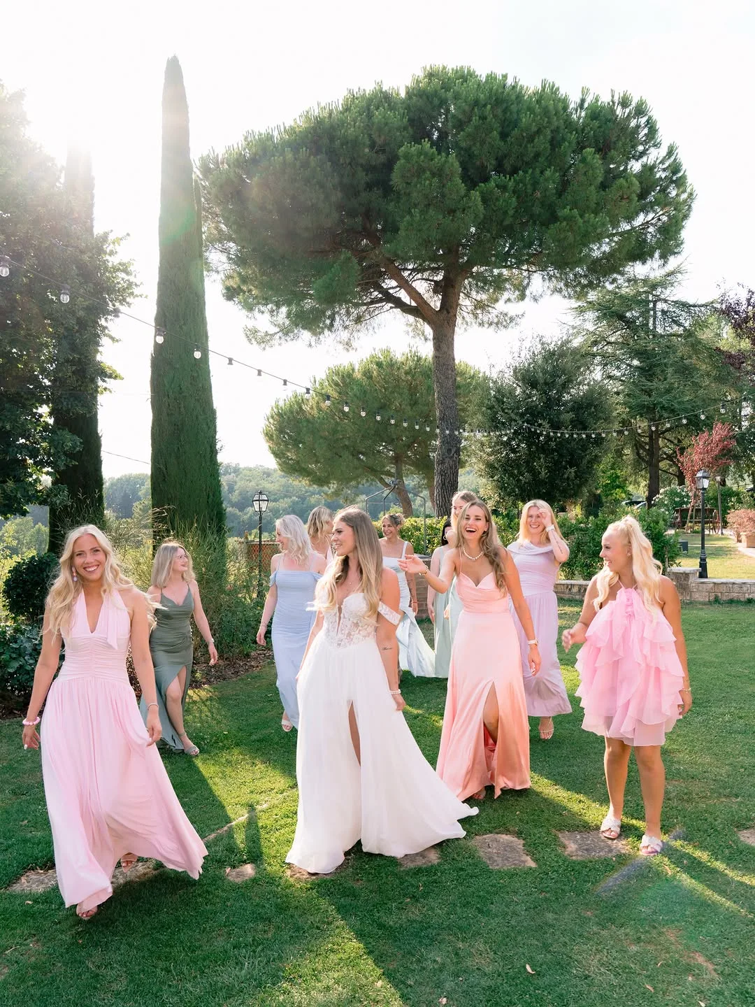 La sposa con le damigelle in abiti pastello in un giardino con pini e cipressi