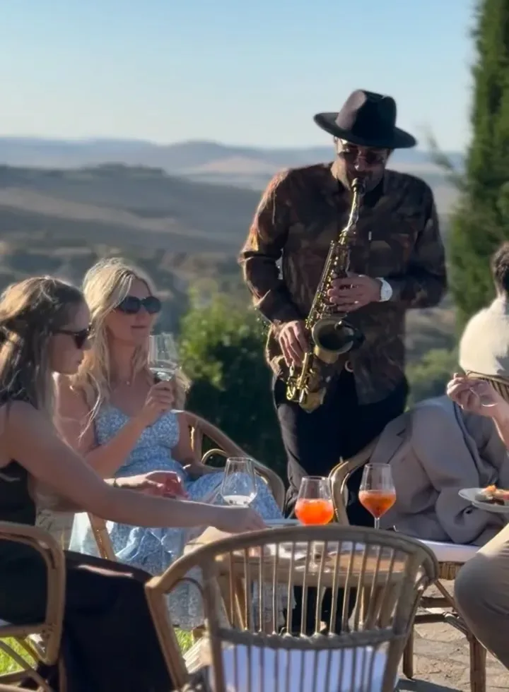 Ciro suona il sax tra i tavoli dell'aperitivo in un giardino con vista sulle colline